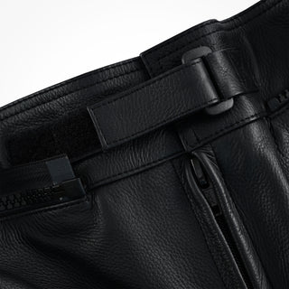 Close-up of the trousers' waist area, showing a leather adjustment strap with a plastic buckle, hook-and-loop (Velcro) fastening, and the top of the main fly zipper.