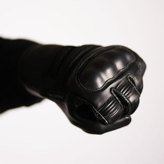 The black leather glove is shown clenched in a riding fist, demonstrating the ergonomic fit and highlighting the solid presence of the hard knuckle protector during flexion.