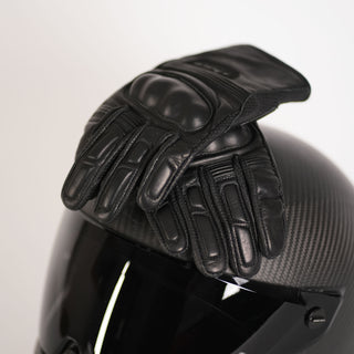 A pair of black leather Apex short cuff motorcycle gloves with hard knuckle protection resting on a dark carbon fiber motorcycle helmet.