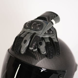 A pair of black and grey Stratus goatskin leather touring gloves with carbon fiber knuckle protection resting on a dark carbon fiber motorcycle helmet.