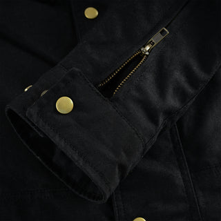 Close-up shot of the jacket's black sleeve cuff, showing the adjustable snap closure flap, a functional brass zipper detail, and the waxed cotton fabric texture.
