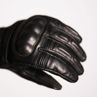 Close-up of the back of the black leather short cuff glove, highlighting the smooth, integrated hard shell knuckle protector and the ribbed finger articulation panels.