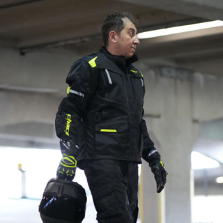Full-body shot of a male rider standing and holding a helmet, wearing the black and hi-vis yellow adventure touring jacket and matching black riding trousers.