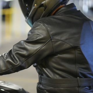 Close-up of the rider's back and shoulder while mounted on a motorcycle, highlighting the quality of the leather, the clean tailoring, and the curved shoulder seams of the black Apex jacket.