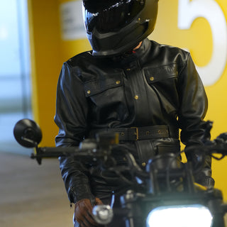 Close-up of a rider wearing the black Apex leather jacket and a carbon helmet, standing near the handlebars of a motorcycle, highlighting the jacket's belted waist and chest pockets.